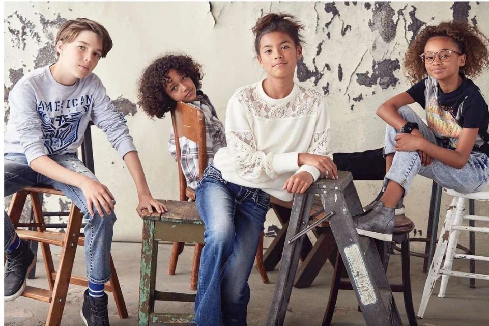 Four children pose indoors wearing casual clothes. One boy sits on wooden ladder next to a girl wearing a white sweater and jeans.