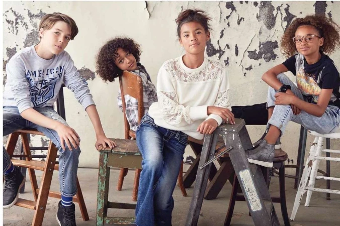 Four children pose indoors wearing casual clothes. One boy sits on wooden ladder next to a girl wearing a white sweater and jeans.