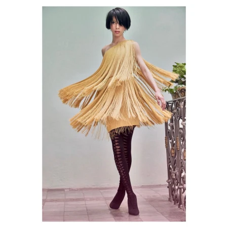 A woman in a one-shoulder dress with fringe poses indoors with a neutral wall and decorative railing, wearing patterned leggings and booties.