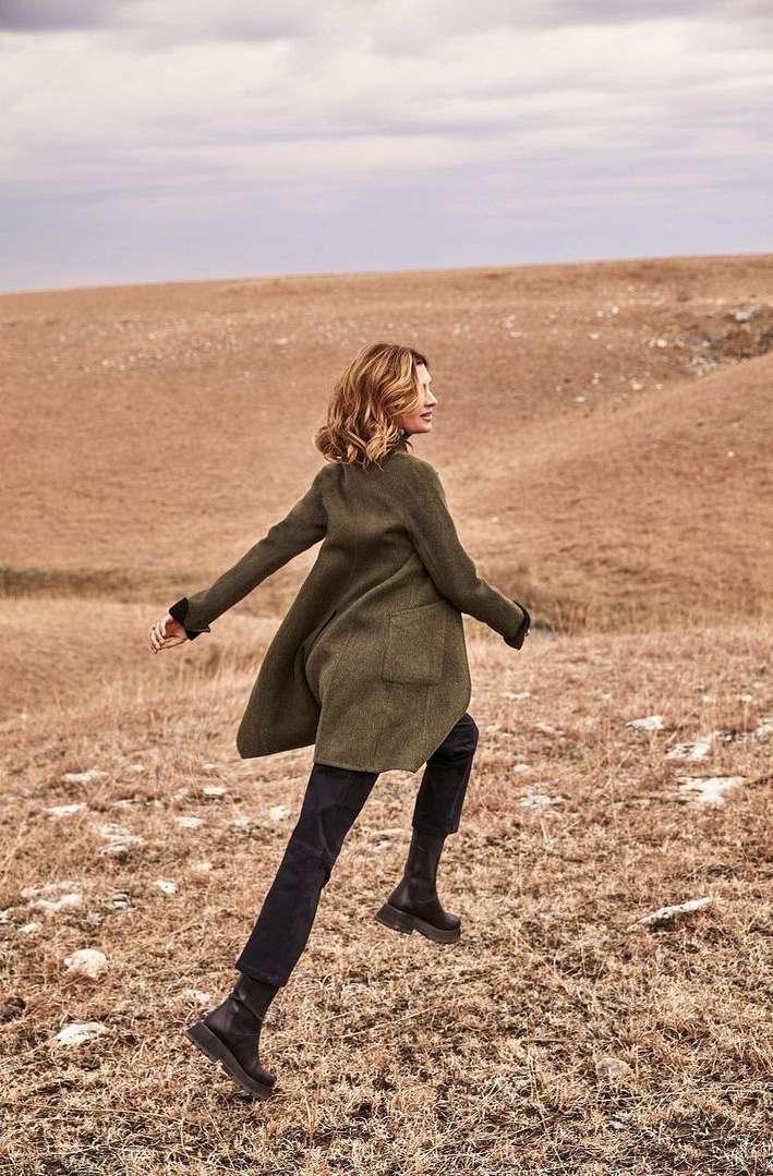 Woman in an olive green coat runs across a dry, hilly, grassy field under a cloudy sky, showcasing outdoor fashion and active lifestyle.