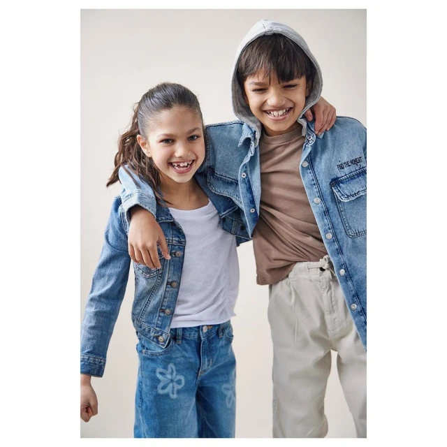 Two smiling children pose indoors against a neutral backdrop. A girl and a boy are wearing casual denim and cotton clothing.