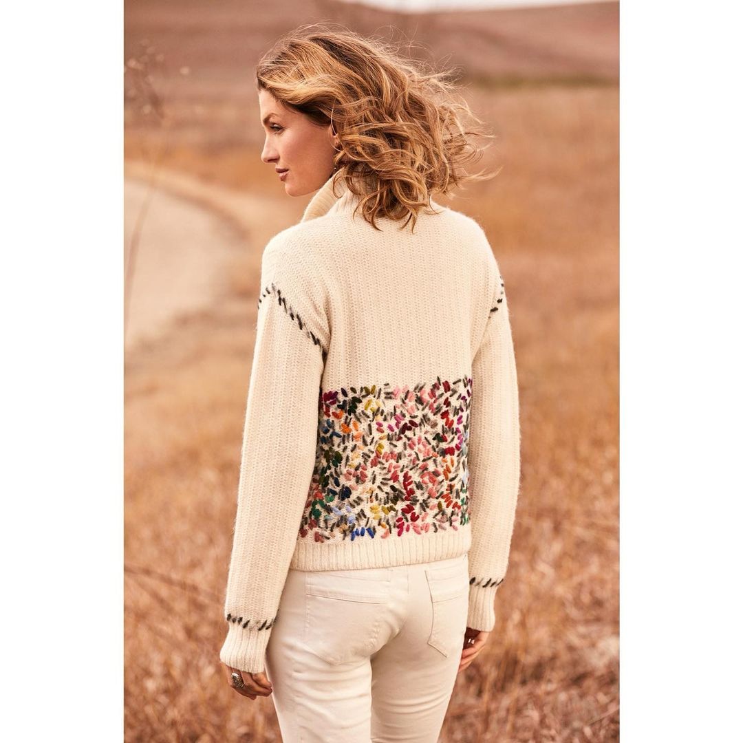 A woman in a cream-colored sweater with floral embroidery stands outdoors, showcasing fashion in a natural light setting with neutral tones.