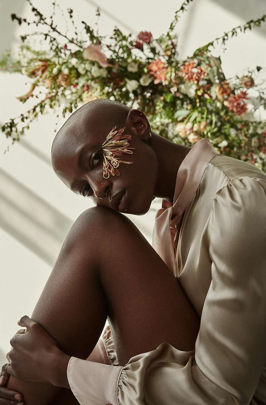 Elegant portrait of a Black woman with shaved head, adorned with floral detail, in a silky beige shirt, complemented by a floral background.