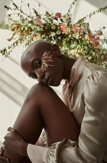 Elegant portrait of a Black woman with shaved head, adorned with floral detail, in a silky beige shirt, complemented by a floral background.