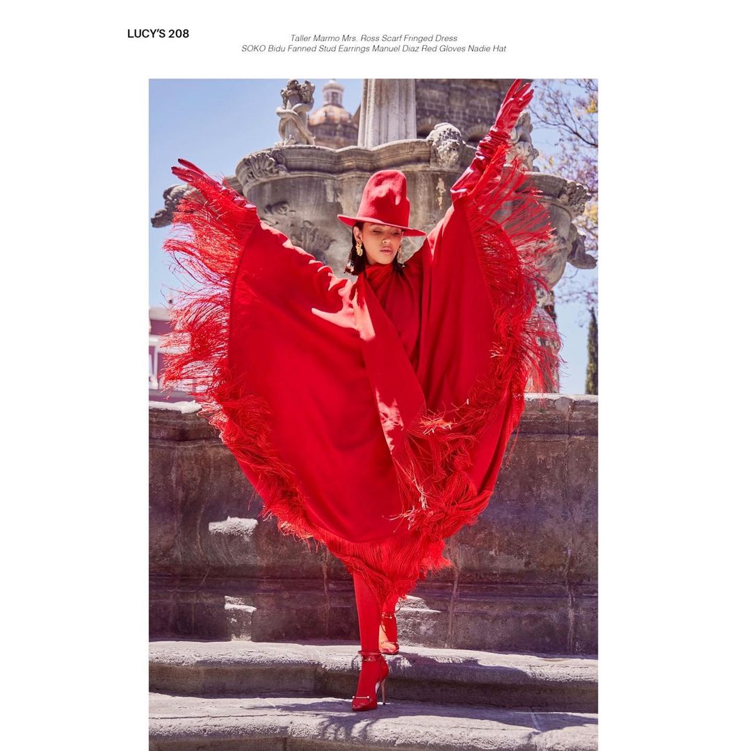 Woman in red hat and fringed cape poses on stone steps in front of a fountain. Fashion editorial in outdoor urban environment.