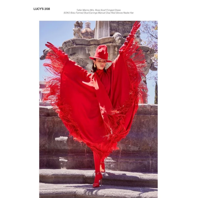 Woman in red hat and fringed cape poses on stone steps in front of a fountain. Fashion editorial in outdoor urban environment.