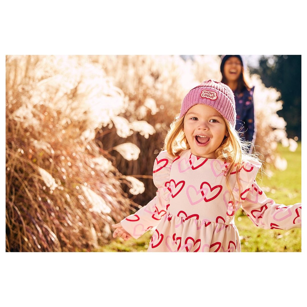 A toddler girl in a heart-patterned dress and pink knit hat smiles in a field with pampas grass, with a blurry young woman in the background.