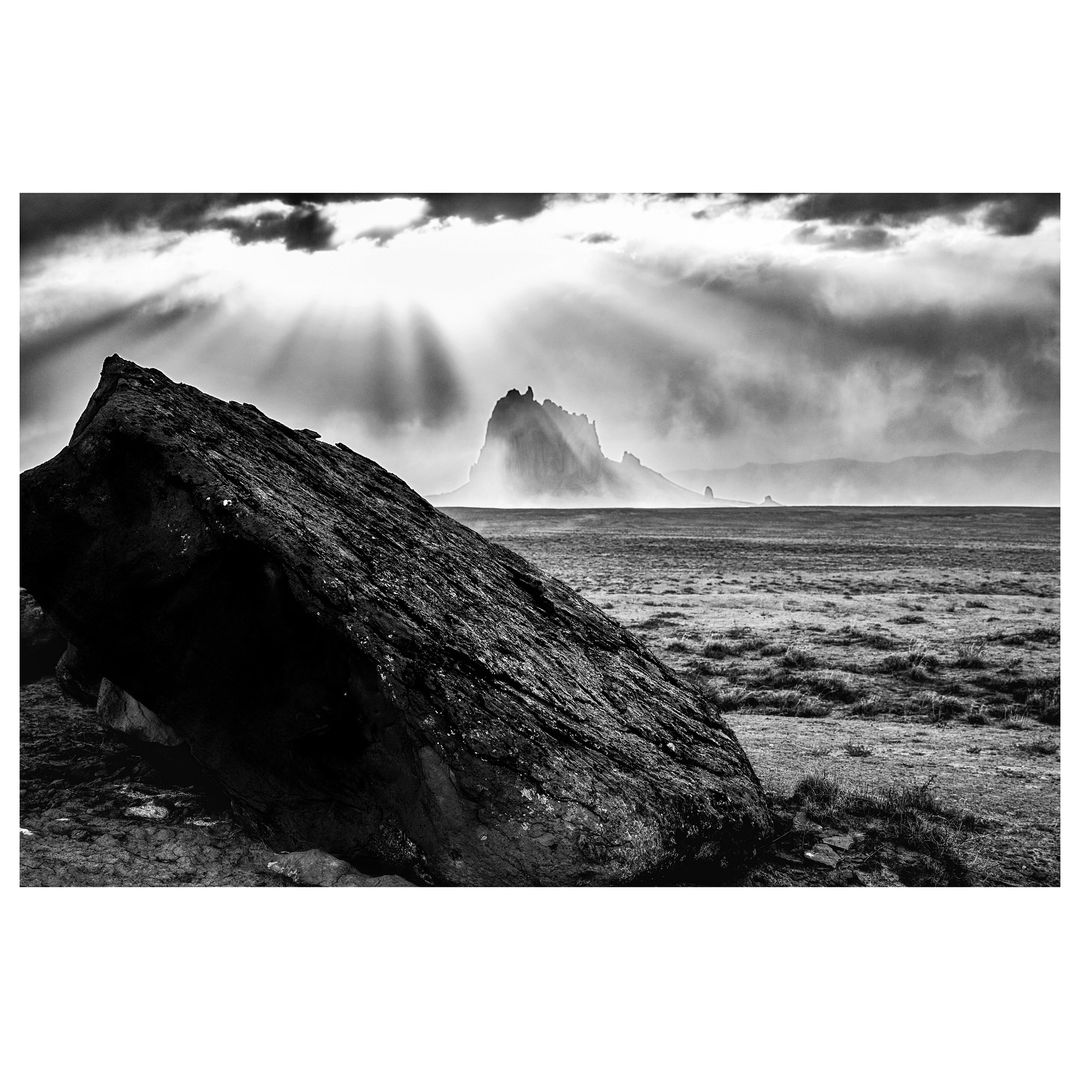 Monochrome landscape with a large rock in the foreground and a distant mountain peak illuminated by sun rays breaking through the clouds.