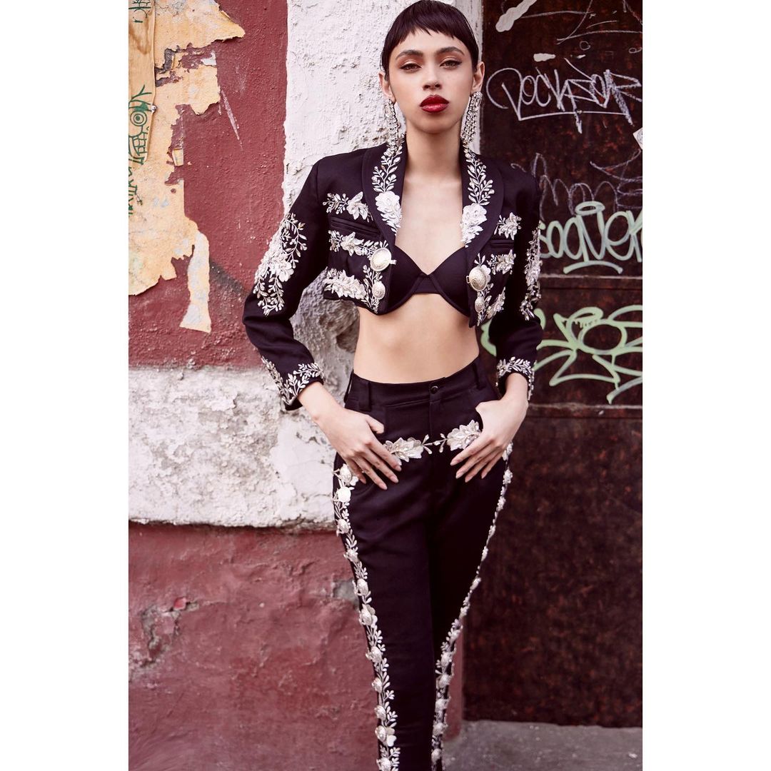A fashionable woman in a black crop jacket and matching pants with white embroidery posing against a textured urban wall with graffiti.