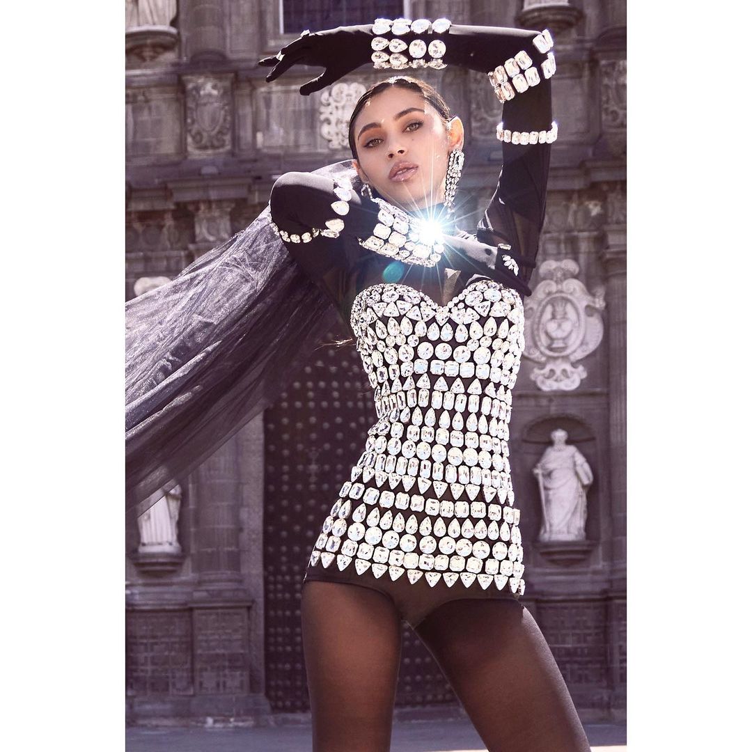 Fashionable woman poses in a sparkling crystal-studded outfit with black tights and gloves, set against a gothic architectural background.