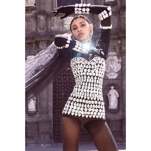 Fashionable woman poses in a sparkling crystal-studded outfit with black tights and gloves, set against a gothic architectural background.