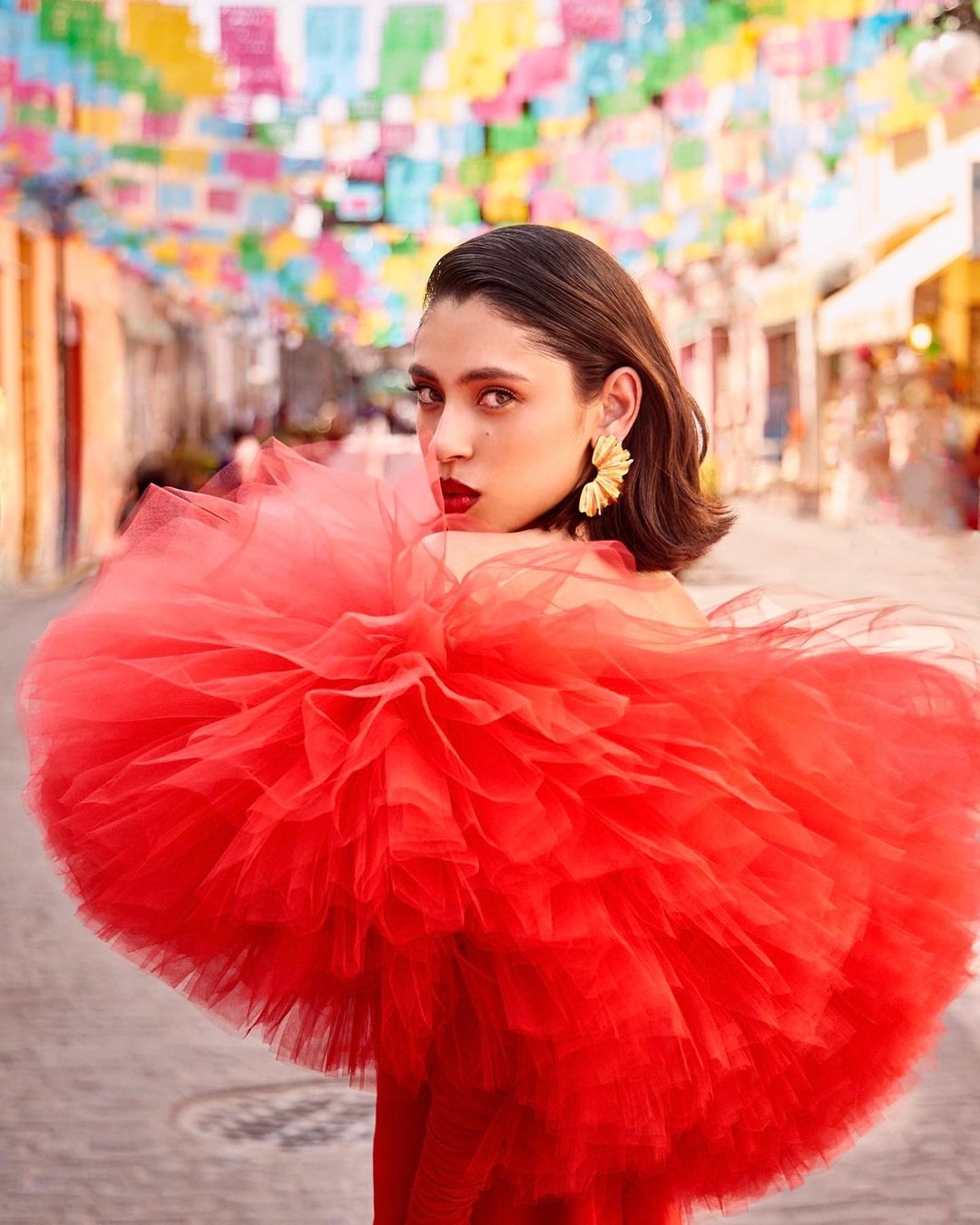 Young woman in red dress posing in street with colorful decorations; medium shot showcases fashion and vibrant environment.