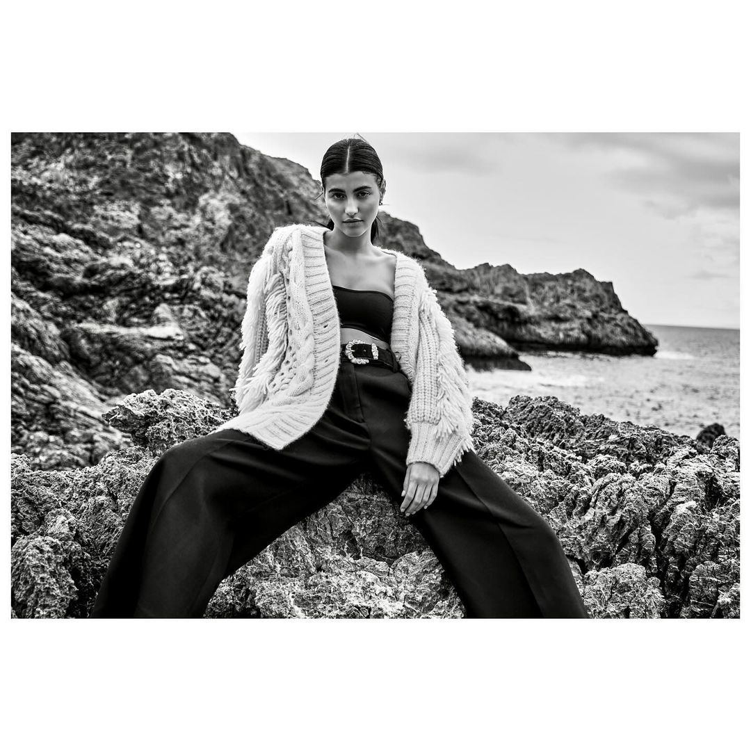 Black and white fashion shot of a woman in a knit cardigan and dark pants sitting on rocks near the sea, looking directly at the camera.