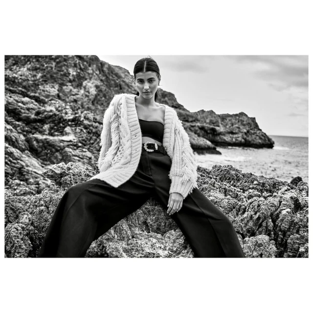 Black and white fashion shot of a woman in a knit cardigan and dark pants sitting on rocks near the sea, looking directly at the camera.