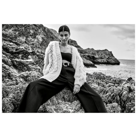 Black and white fashion shot of a woman in a knit cardigan and dark pants sitting on rocks near the sea, looking directly at the camera.