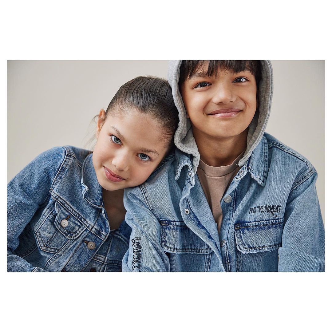 Portrait of a smiling boy and girl in light denim jackets, posing closely together against a neutral background, looking at the camera.