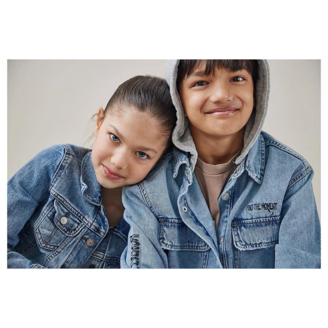 Portrait of a smiling boy and girl in light denim jackets, posing closely together against a neutral background, looking at the camera.