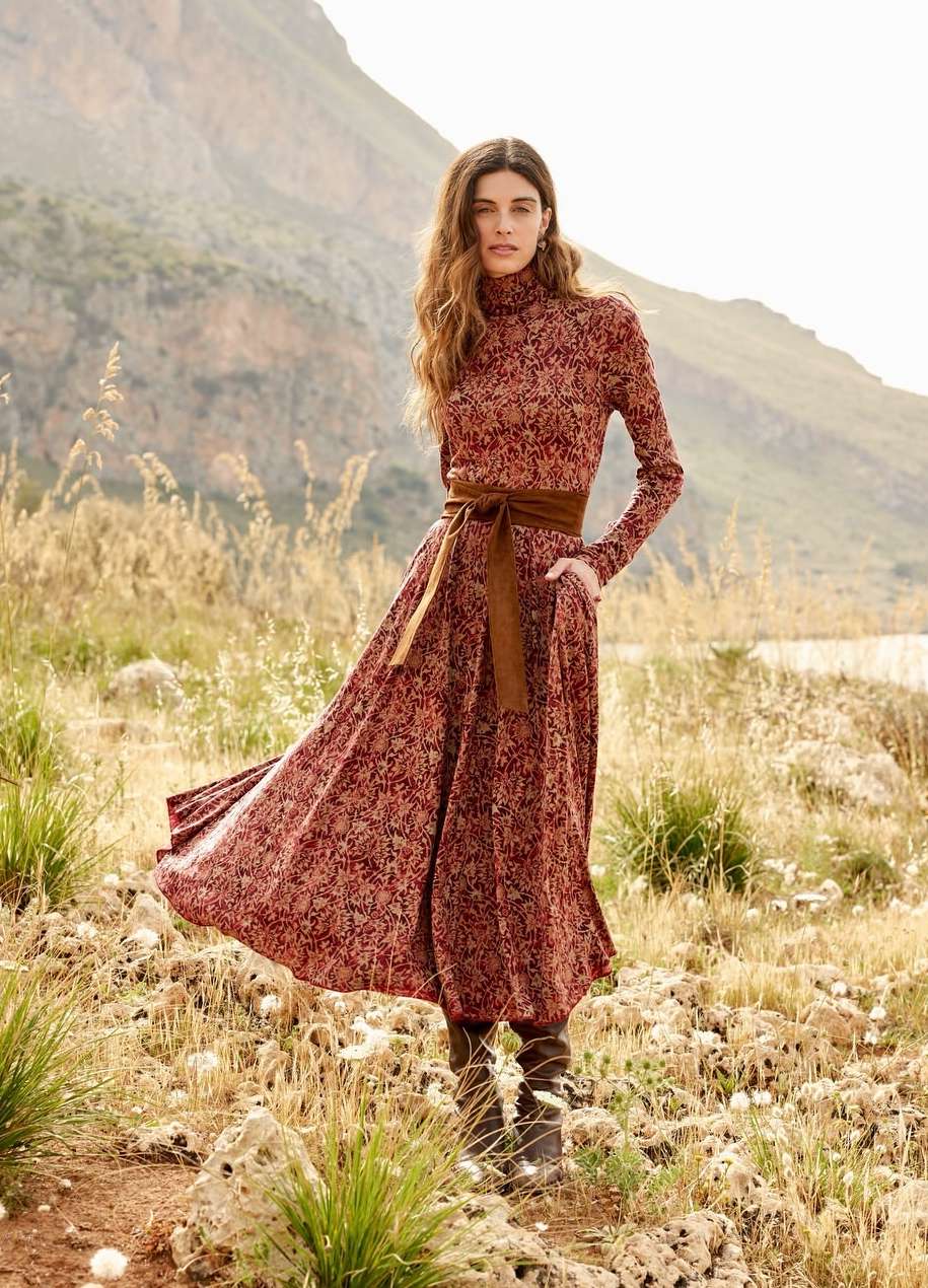 Woman standing in a meadow wearing a patterned red turtleneck dress and brown belt with mountains behind
