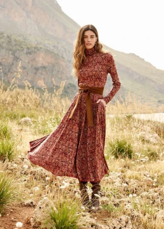 Woman standing in a meadow wearing a patterned red turtleneck dress and brown belt with mountains behind