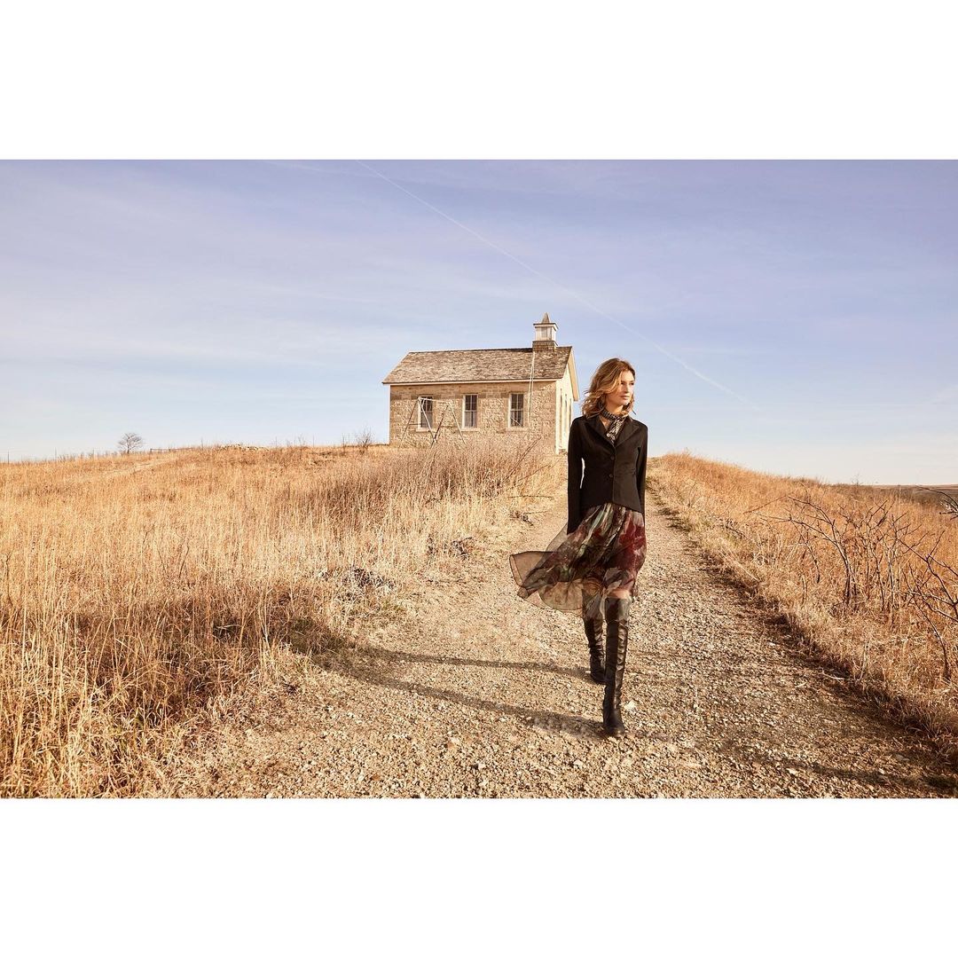 A woman in a dark jacket and floral dress walks along a dirt path towards the camera, with a prairie and stone building in the background.