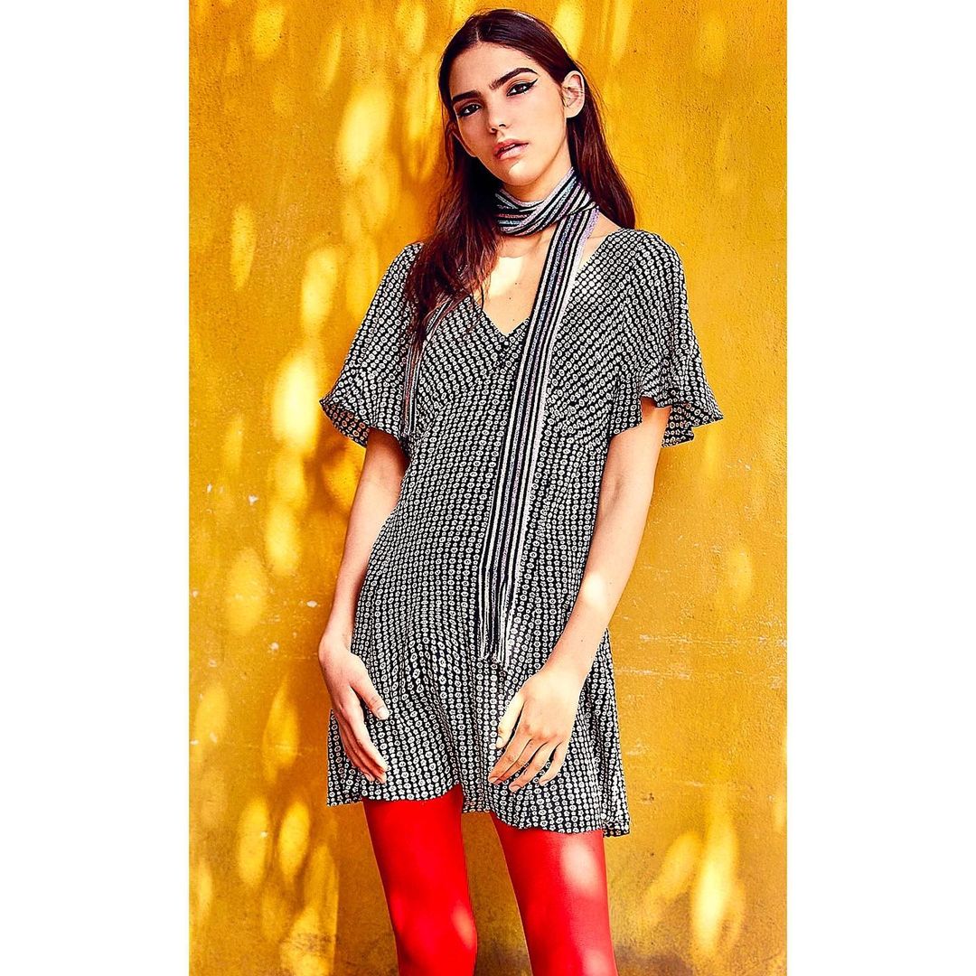 Woman in patterned mini dress and red tights posing against a yellow wall with sunlight creating shadows, fashionable and striking.