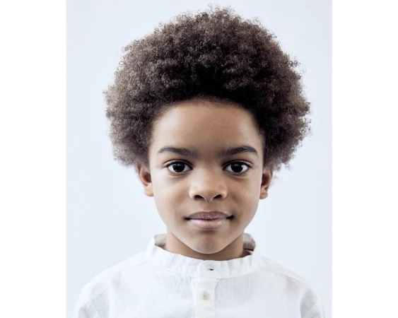 Portrait of a smiling young African-American boy with an afro, wearing a white linen shirt against a plain light blue background.