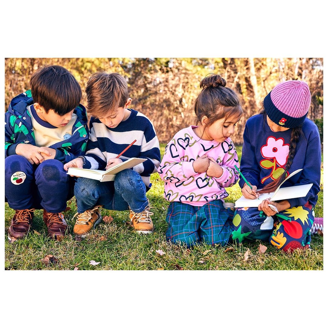 Four children are outdoors sitting in the grass. They are drawing and coloring in notebooks on a sunny day in nature.