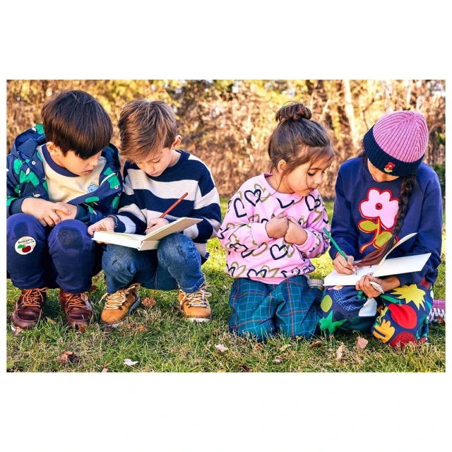 Four children are outdoors sitting in the grass. They are drawing and coloring in notebooks on a sunny day in nature.