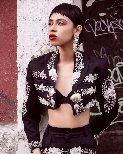 A young woman poses wearing a decorative black cropped jacket with elaborate white embroidery, statement earrings, and bold red lipstick.
