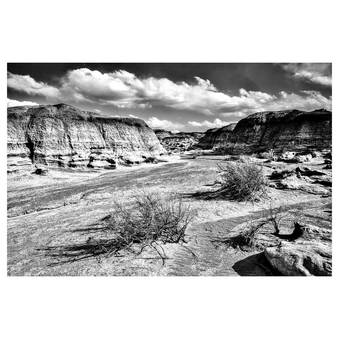Monochromatic landscape featuring eroded rock formations, sparse vegetation, and dramatic sky with clouds casting shadows on the terrain.