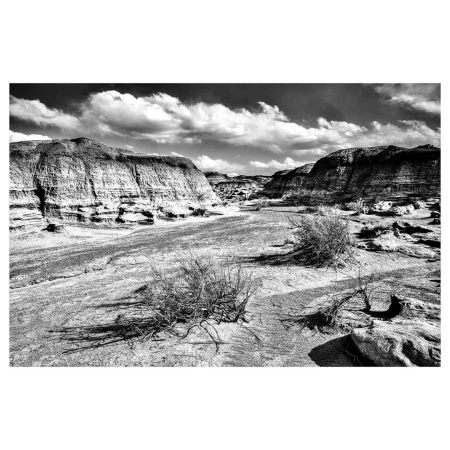 Monochromatic landscape featuring eroded rock formations, sparse vegetation, and dramatic sky with clouds casting shadows on the terrain.