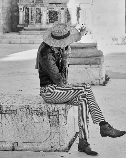 Black and white photo of an adult wearing a hat and striped pants sitting on a stone block outdoors with a building in the background.