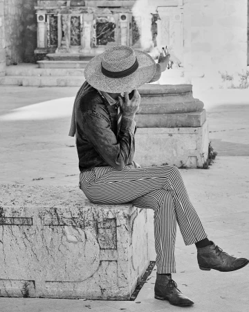 Black and white photo of an adult wearing a hat and striped pants sitting on a stone block outdoors with a building in the background.