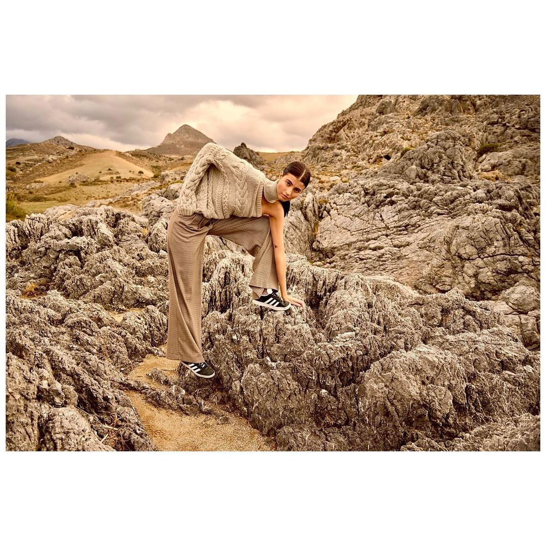 A brunette woman in a beige knit sweater and brown plaid pants poses on rocky terrain, showcasing fashion apparel in a natural outdoor setting.