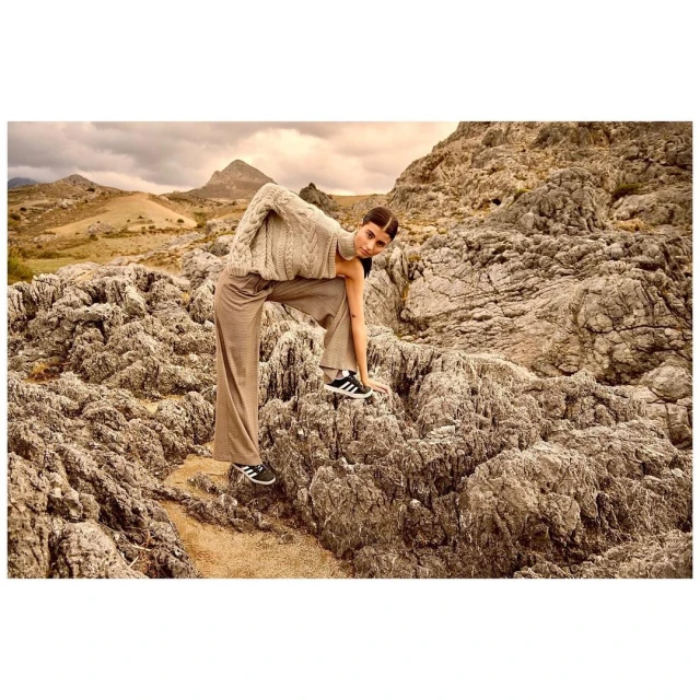 A brunette woman in a beige knit sweater and brown plaid pants poses on rocky terrain, showcasing fashion apparel in a natural outdoor setting.