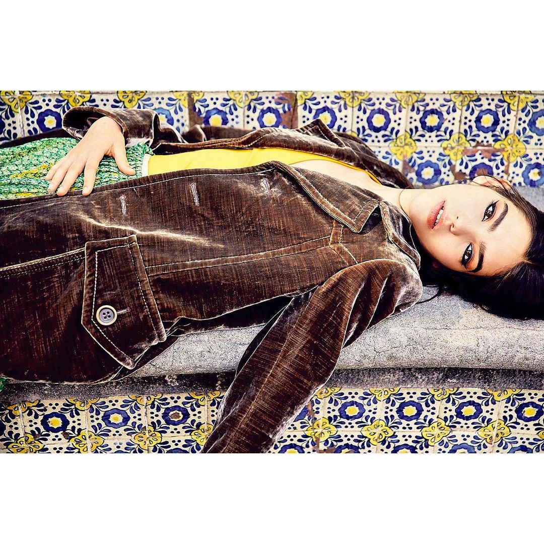 Young woman with dark hair lies on a decorated bench wearing a brown jacket and yellow top, looking up towards the camera.
