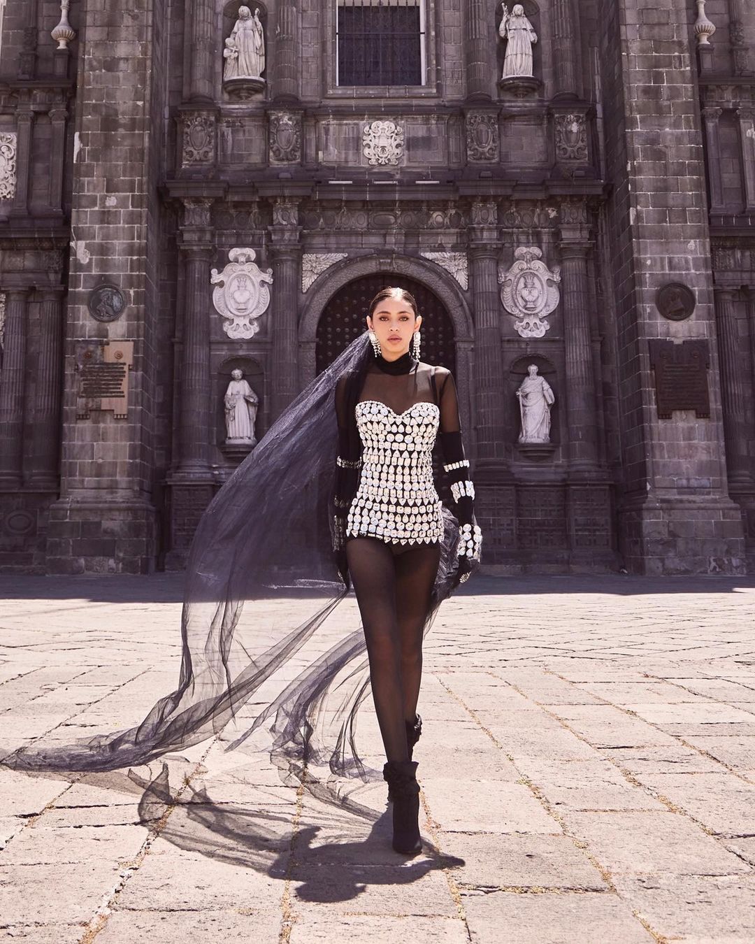 Woman in sheer dress with embellishments and flowing veil poses in front of a historic stone cathedral on a sunny day.