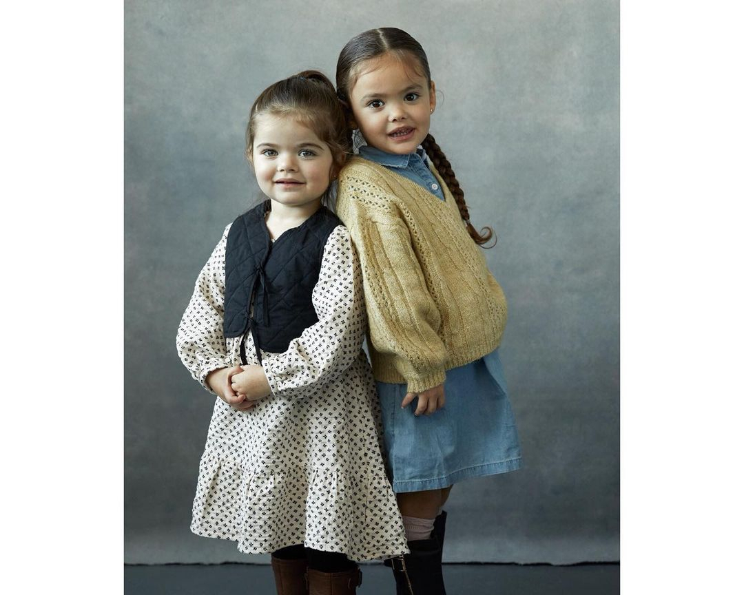 Two young girls in a light grey studio setting. One wears a floral dress with a black vest, and the other wears a denim dress with a knit sweater.