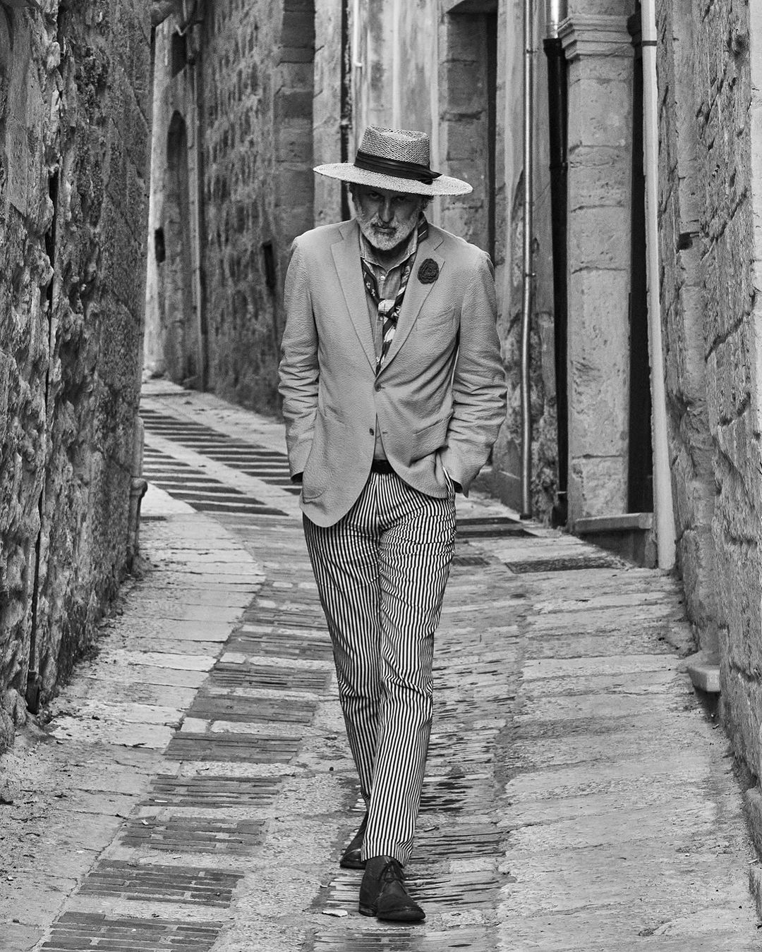 Stylish older man in a hat and suit walks along a narrow stone alleyway. Black and white fashion shot with a European background.