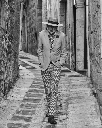 Stylish older man in a hat and suit walks along a narrow stone alleyway. Black and white fashion shot with a European background.