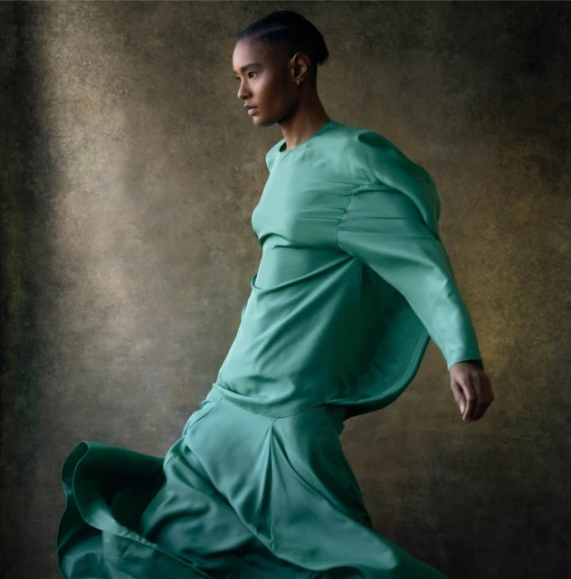 A woman in a flowing teal dress is captured in motion against a muted brown background in a fashion-style studio portrait.