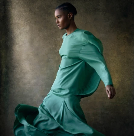 A woman in a flowing teal dress is captured in motion against a muted brown background in a fashion-style studio portrait.