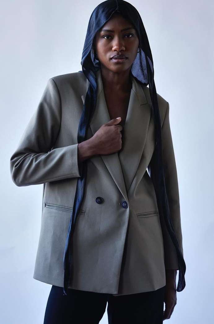 Fashion portrait of Black woman wearing an olive green blazer and a black headscarf against a white backdrop in a studio.