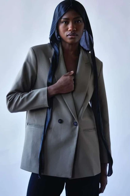 Fashion portrait of Black woman wearing an olive green blazer and a black headscarf against a white backdrop in a studio.