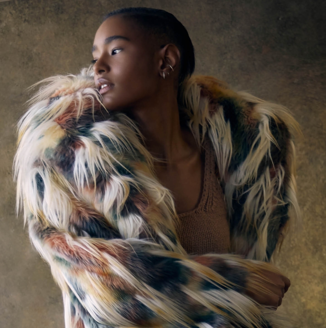 Portrait of a Black woman in a multicolored faux fur coat, looking to the side against a diffused brown background in a studio setting.