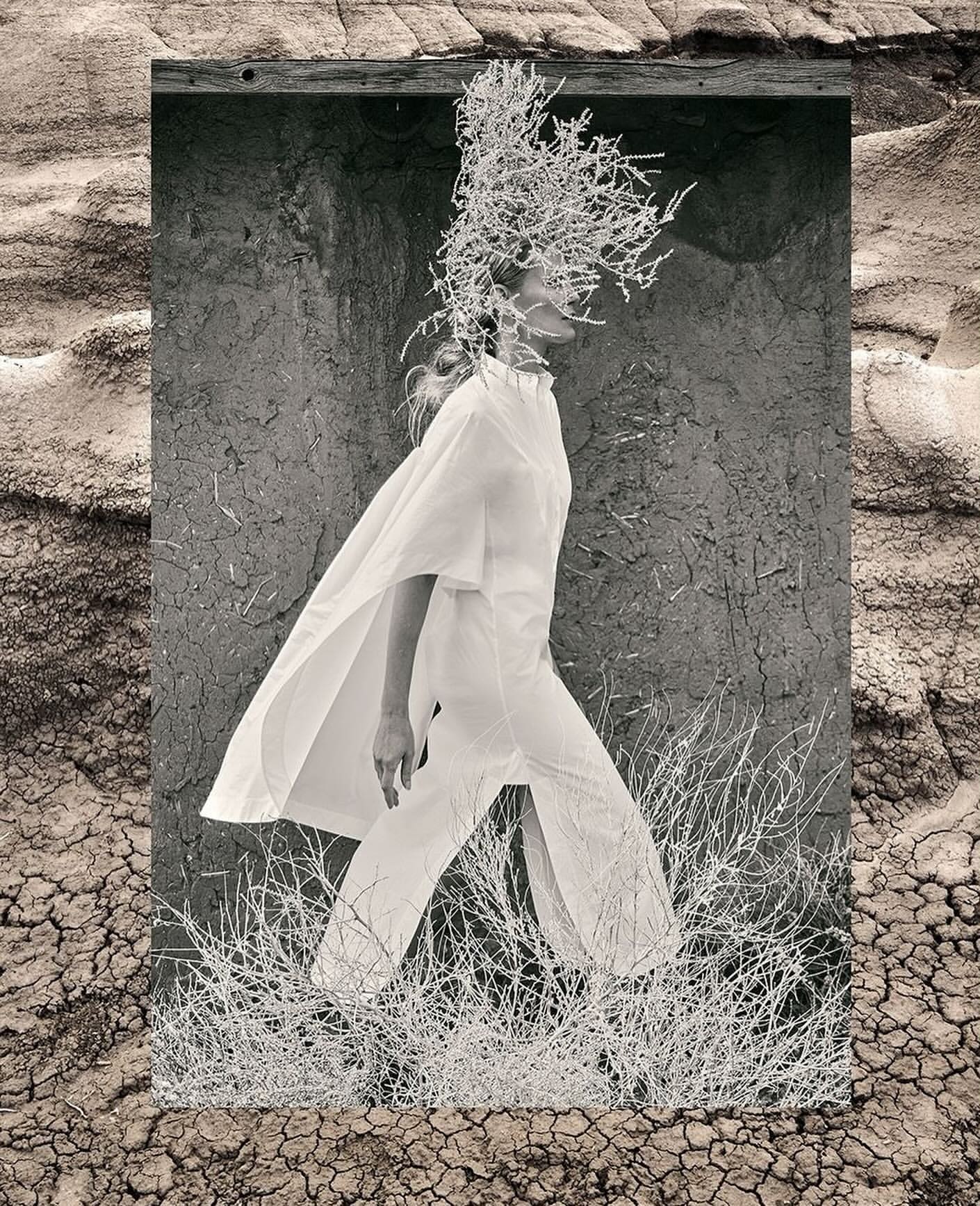 Monochrome fashion shot featuring a woman in flowing white clothing walking through a surreal desert landscape with cracked earth and sparse vegetation.