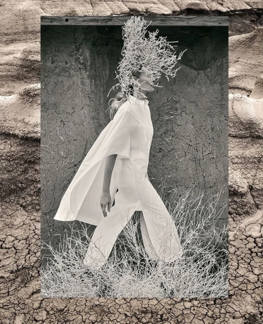 Monochrome fashion shot featuring a woman in flowing white clothing walking through a surreal desert landscape with cracked earth and sparse vegetation.