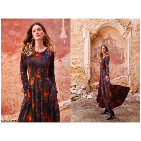 A woman models a long-sleeved, floral-print dress outdoors, against weathered stucco walls in a warm color palette.