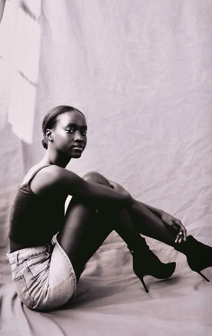 Studio shot of a Black woman sitting cross-legged wearing a tank top, denim skirt, and high heel boots against a muslin backdrop.