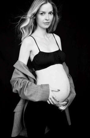 Black and white portrait of a pregnant woman in a bra and blazer, posing indoors against a black backdrop, emphasizing maternity and fashion.